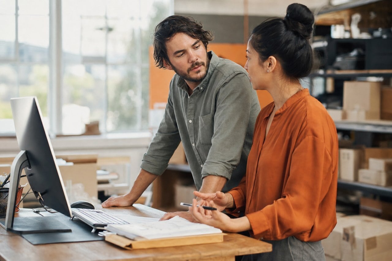 A customer service rep and a finance colleague problem-solving together at a standing desk in a bright distribution office.