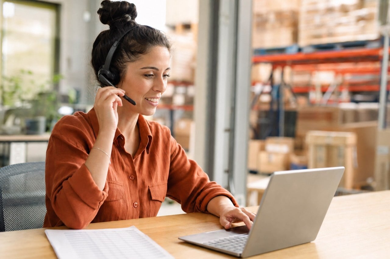 A customer service rep wearing a headset confirming an order on a laptop at an order desk in a bright distribution office.